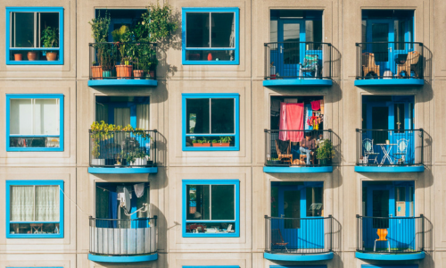Beige apartment building with blue window frames and balconies filled with green plants.